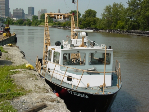 A black & white buoy tender boat, docked in a canal in front of the Buffalo skyline. It has an A-frame boom