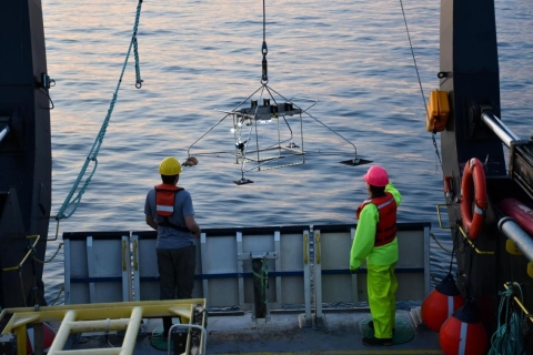 Two people on a large boat guide an equipment frame over the side of the boat as it is lifted by a crane