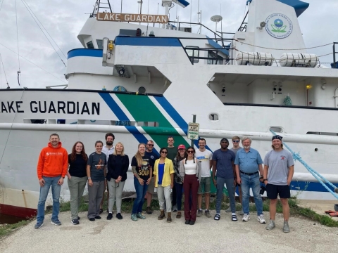 Sixteen people pose for a picture in front of a large ship "Lake Guardian"