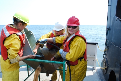 Three people work on the deck of a boat, wearing hard hats, life jackets, and rain gear. One is pouring a tub of mud into a basin on a platform while the other two move the mud into the basin with hands and water from a hose.