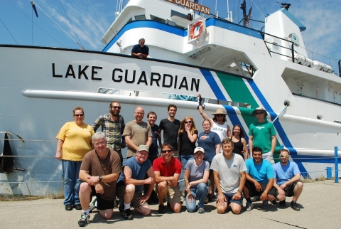 A group of 17 people pose for a picture in front of a large boat named "Lake Guardian." Another person is up on the deck of the boat.