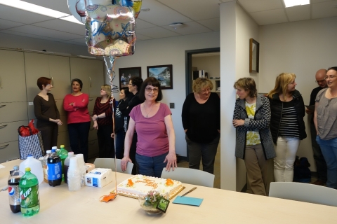 A person stands next to a balloon, cake, and plant, while guests stand in the background.