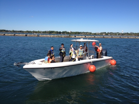 A white boat in the water near shore with several people in life jackets aboard. One waves to the camera. There are round fenders hanging over the sides.