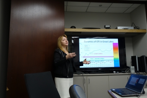 A person with long hair standing next to a screen giving a presentation.