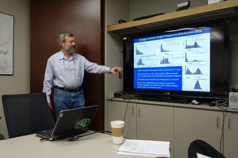 A person with short gray hair and a beard standing next to a screen giving a presentation.