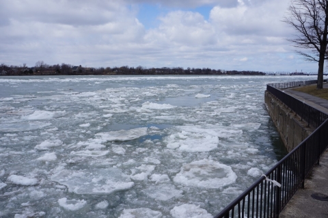 Ice floes in a river near a bend in a park with vertical concrete bulkheads and a metal fence