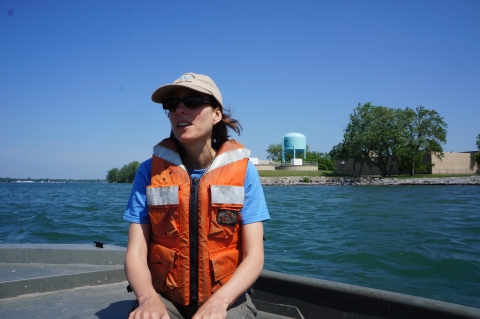 A person wearing a life jacket sits in the front of a boat on the water. There is a water tower on the land behind them.