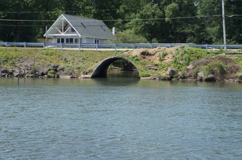A large culvert runs under a roadway, connecting a stream with a larger body of water. There is a home on the far side of the roadway.