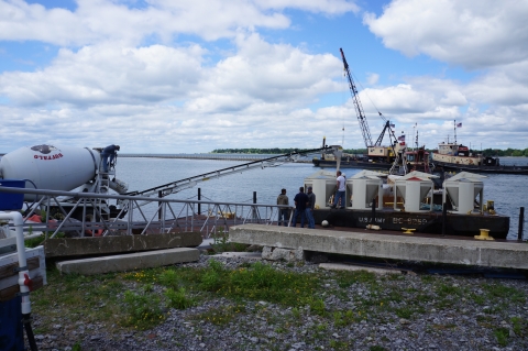 A cement truck pours cement into containers on a barge at the waterfront. In the background is a tug and a barge with a crane.