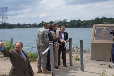 Four people stand on a concrete walkway near the water, cutting a ribbon. A fifth person stands nearby, giving a thumbs up.