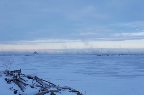 Snow and ice cover a body of water. A breakwall contains the scene, with fog rising from just beyond the breakwall.