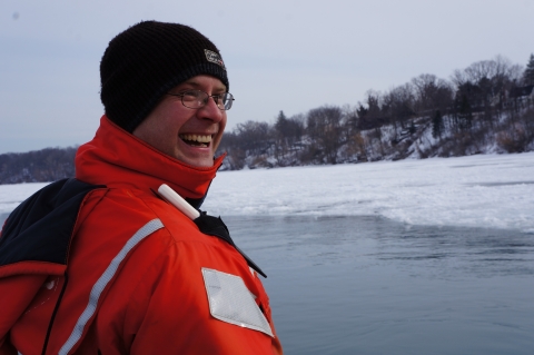 A person in an orange float suit and winter hat smiles as they stand by an icy river.
