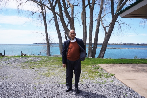 A bald person with glasses in a suit jacket over a sweater standing outside at the waterfront