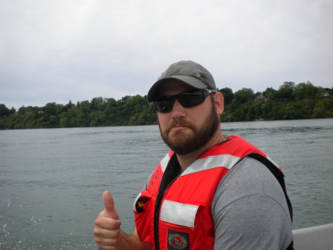 A bearded person in a baseball hat, sunglasses, and a life jacket giving a thumbs up while on a boat.