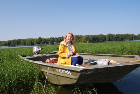 A person in a raincoat sits in a small boat that is surrounded by reeds in a lake.