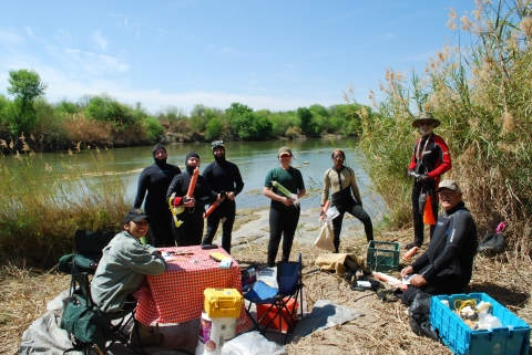 Several people in wetsuits stand by the edge of the river. There is a table set up and the divers are holding equipment, getting ready to go dive.