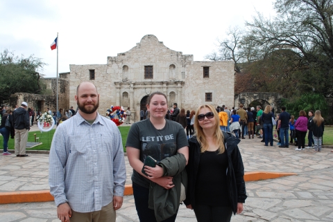 Three people pose for a picture in front of a historic building. There are crowds of people nearby and a green with a flagpole with a Texas flag and memorial wreaths.