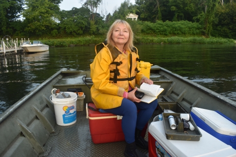 A blond woman sits on a cooler in a flat-bottomed boat near shore, there are buckets and coolers around her and a notebook in her hands