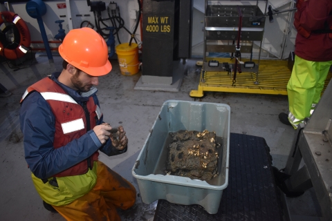 A person in a life jacket and hard hat kneels on the deck of a boat next to a tub of mud and dreissenid mussels to take a sample for the jar in their hands.