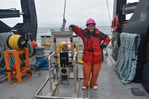 A person in raingear stands on the deck of a large boat and leans their arm on a large frame holding a camera and other instruments.