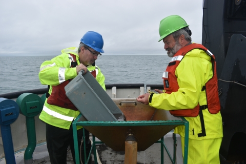 Two people wearing raingear and life jackets work on the deck of a large boat. They are standing at a basin while one empties a tub of mud and water into it.