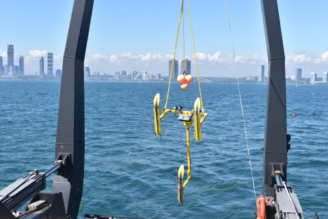 A metal frame with three skis is lowered down toward the water using a large metal A-frame and winch. A city is on the horizon.