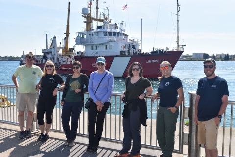 Seven people pose at a railing by the water. A large boat named LIMNOS is in the water behind them.