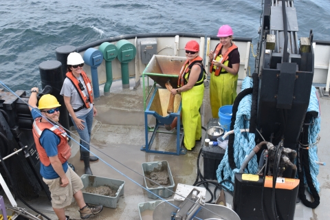 Four people wearing hard hats and life jackets work on the deck of a large boat. There are a few tubs of mud on the deck and a wash table setup.