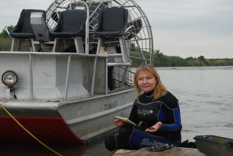 A person in a wetsuit sits on a rock next to a metal boat with a large fan on the back of it. The person is holding a notebook and has a pile of mussels on the rock next to her