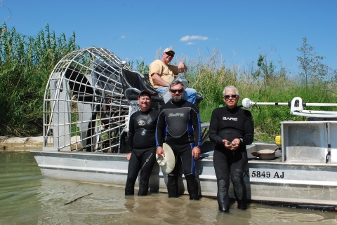 Three people in wetsuits stand in shallow water next to a metal boat with a large fan on it. A person is sitting on the boat above the other three.
