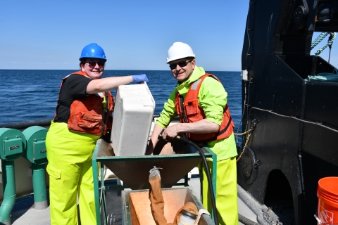 Two smiling people in safety gear work at a basin on the deck of a large boat. One person dumps a tub into the basin while the other washes the tub with a hose