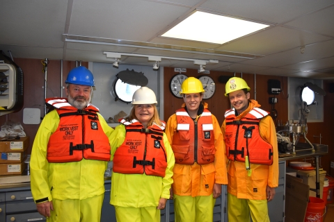 Four people wearing rain gear, hard hats, and life jackets indoors on a boat. There are portholes in the background, gear, and two clocks labeled "Ship Time" and "UTC ZULU."