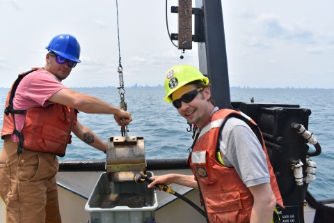 Two people working on a boat on a lake, wearing hardhats and life jackets. One holds a metal scoop with mud over a tub while the other washes it out.