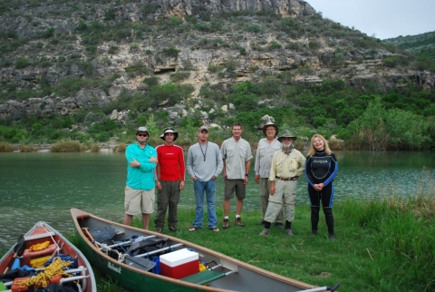 Seven people stand together by a river and some canoes. The terrain behind them is rugged