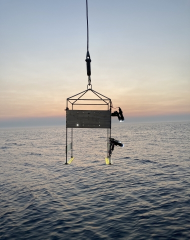 A metal frame being suspended above the water at sunset. The frame has several lights attached and runners with ruler demarcations on the base.