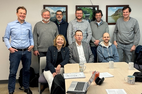 Nine people pose for a group photo in an office, six people standing behind three people seated at the end of a table