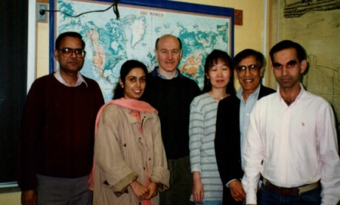 a group photo with three medium brown-skinned men, a medium-brown skinned woman, a light-skinned woman, and a light-skinned man in front of a world map in a classroom