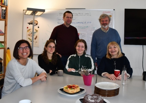 Six people pose for a picture at a small party in a meeting room. Four people sit and two stand behind them. There are cakes and dishes on the table