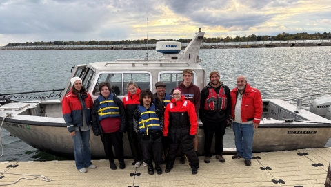 Nine people in float coats or life jackets stand on a dock by a boat in a river