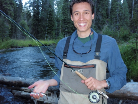 A person with light medium-toned skin and short dark hair smiles while holding a fishing rod and a small fish while standing in a stream in a conifer forest.