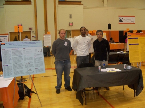 Two graduate students and a researcher stand behind a table next to academic posters on easels in a gymnasium