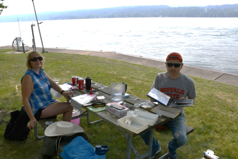 Two people sit at a picnic table by the water doing biological sampling work. There are trays of samples and supplies on the table.