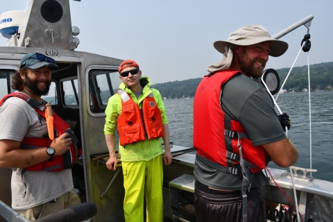 Three people working on the deck of a metal boat with a cabin. One is holding a rope on a winch and pulley going over the side of the boat, and everyone has turned to smile at the camera.