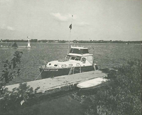 Black and white picture of a boat with a cabin tied up to a dock in a river.