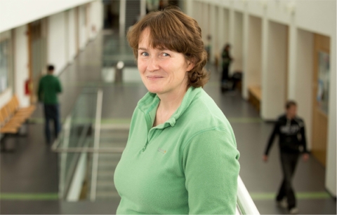 A person with light brown shoulder length hair with fringe posing in the atrium of an academic building. A few people are walking near the classrooms in the background.
