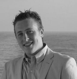 Black and white portrait, a young person with short styled hair smiles while wearing a light suit with open collared shirt in front of the sea