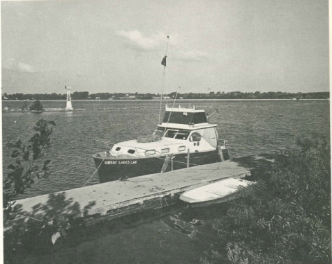 black and white photo of a mid-sized boat with cabin at a dock in a river