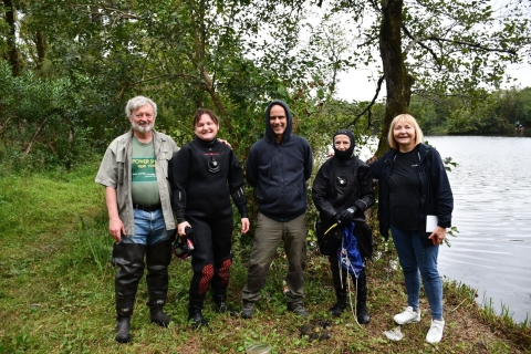Group photo of five people posing by trees by the water on an overcast day. Two people are wearing wet suits