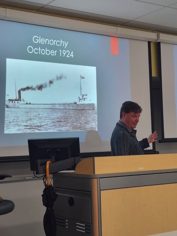 A person lecturing at the front of a room standing in front of a projector screen that says "Glenorchy October 1924" with a black-and-white photo of a freighter ship
