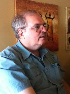 A person with short gray hair and moustache and large wire-frame glasses looks out of frame, wearing a blue short-sleeve button-up shirt, sitting in a room near some paintings